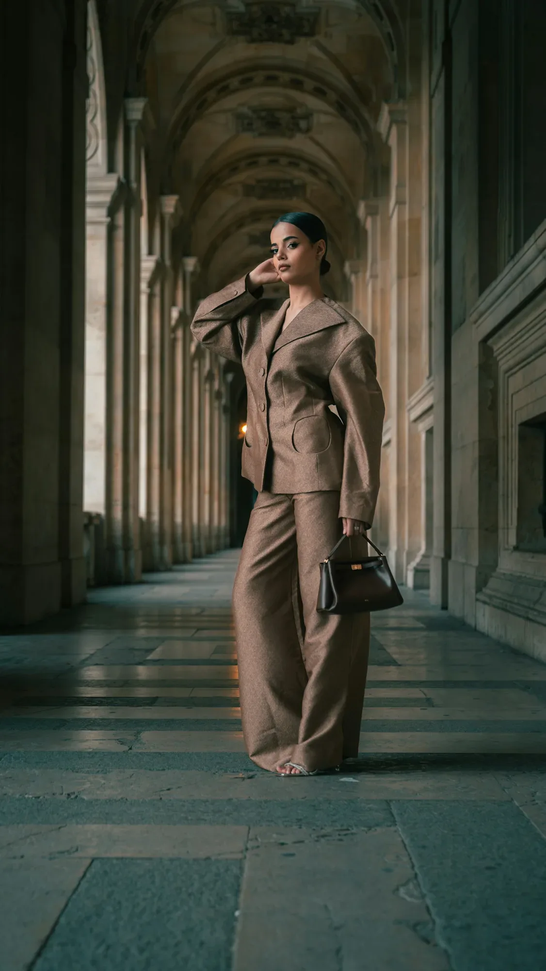 Une femme en tailleur beige, un sac à la main, sous une galerie de pierre